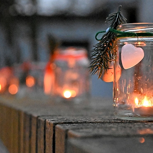 Ein mit Kerze beleuchtetes Glas ist mit Herz und Tannenzweig dekoriert, weitere Gläser im Hintergrund.