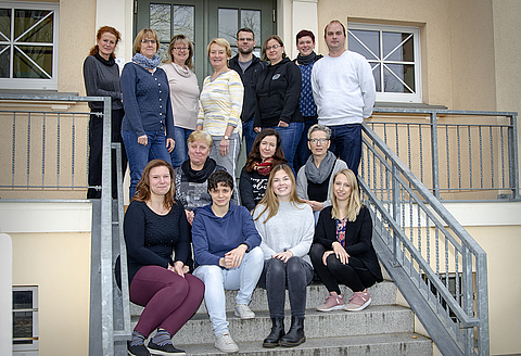 Gruppenfoto: Das Team des Kindergartens steht und sitzt auf der Treppe des Hauses zum Garten und lächelt in die Kamera.
