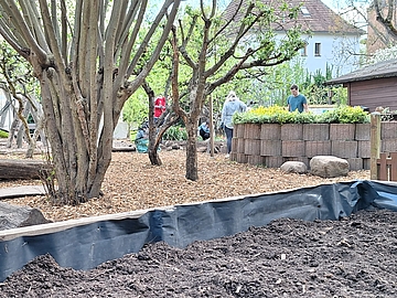 Eltern und Kinder arbeiten im Kitagarten. Im Vordergrund ist ein mit Erde gefülltes Beet zu sehen, im Hintergrund pflegen Helfende die Beete und verteilen Hackschnitzel.