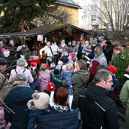 Viele Kinder und Erwachsene singen zusammen im Garten des Kindergartens vor einer geschmückten Holzhütte.