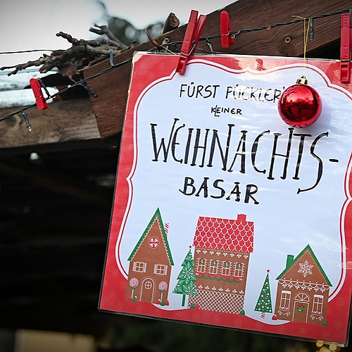 Ein Schild mit der Aufschrift "Weihnachtsbasar" hängt mit roten Kugeln dekoriert an einer Holzhütte.